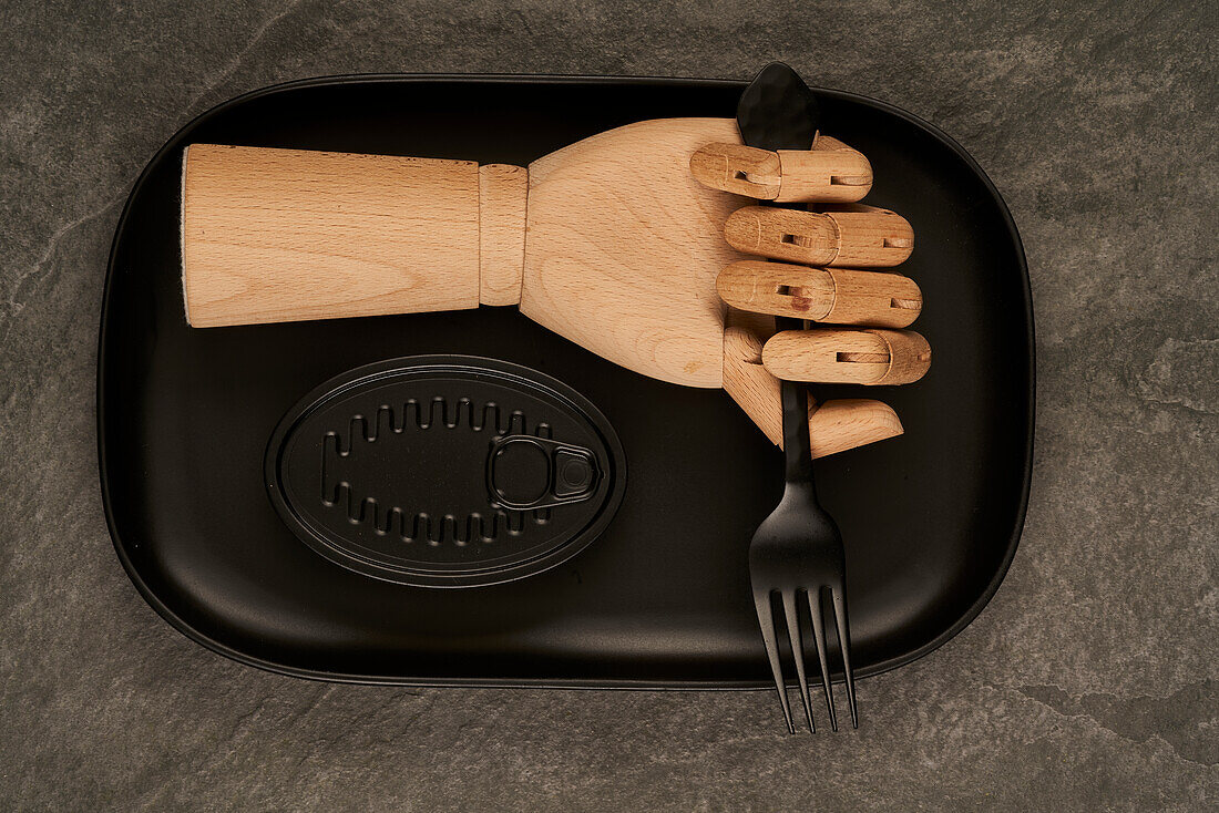 Künstliche Holzhand mit Gabel auf Tablett neben versiegelter schwarzer Dose mit Konserven auf Tisch