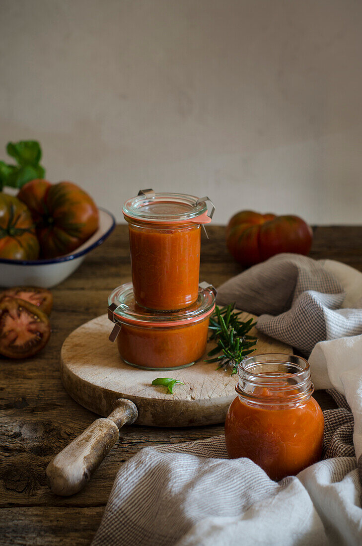 Full glass jars with homemade natural fried tomato sauce placed on cutting board board near with fresh green rosemary and basil leaves placed on rustic wooden table