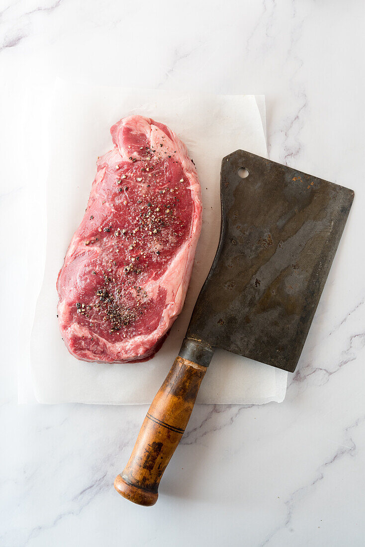 Overhead view of uncooked meat piece with black pepper against baking paper and hatchet knife on marble background