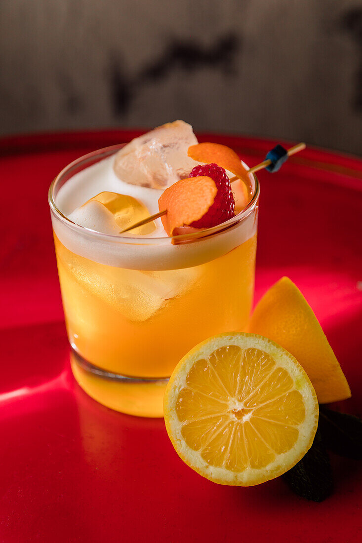 Crystal glass of Amaretto Sour cocktail garnished with orange zest and raspberry served with halved lemon