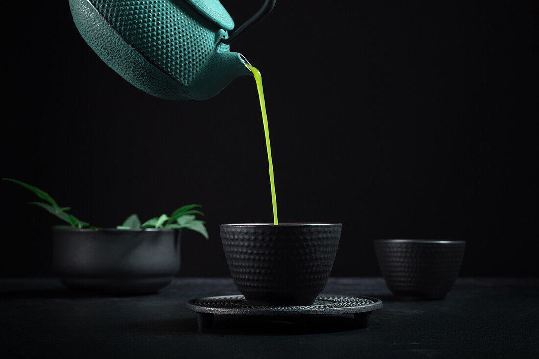 Gesunder japanischer Matcha-Tee, der während einer Teezeremonie aus einer grünen Teekanne in eine schwarze Keramikschale gegossen wird, vor schwarzem Hintergrund