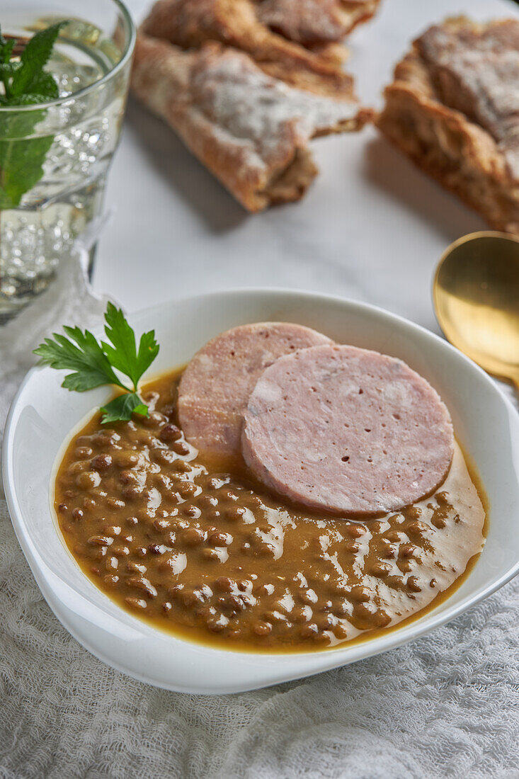 From above spoon placed near bowl of yummy lentil soup served with slices of sausage and leaf of parsley on marble table and gray napkin