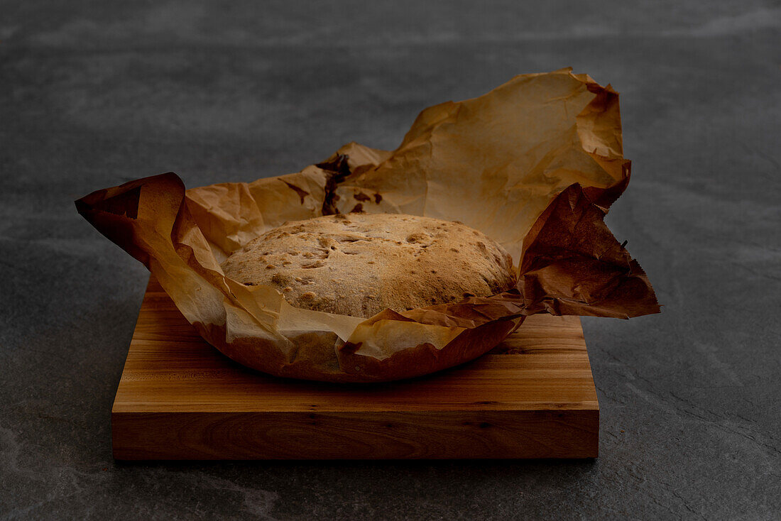 Frisch gebackenes, handwerklich hergestelltes, rundes Brot mit knuspriger Kruste auf Pergamentpapier vor einem schwarzen Hintergrund