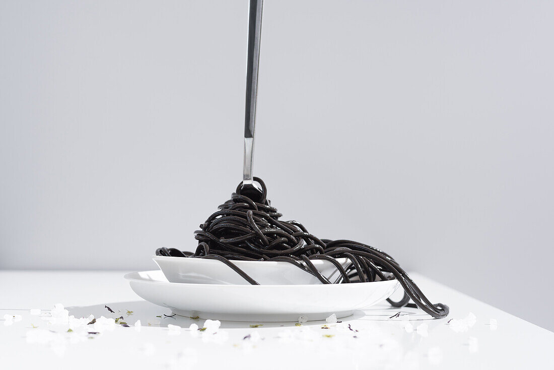 Stainless fork in full bowl of black spaghetti with cuttlefish ink on white table with thin blooms in studio on gray background