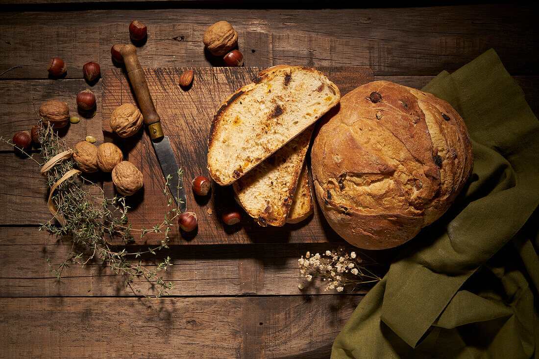 Draufsicht auf ein köstliches hausgemachtes Sauerteigbrot mit Nüssen auf einem Holzbrett mit Messer und Kräutern auf einem rustikalen Tisch