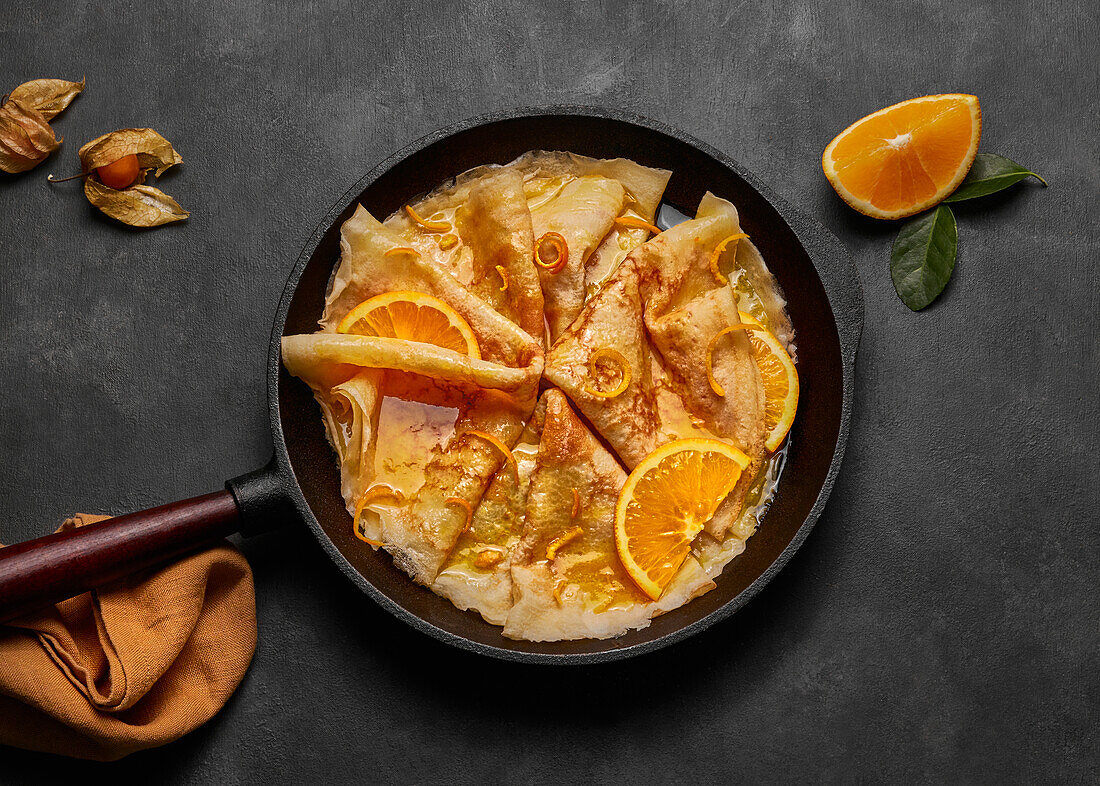 From above homemade crepes suzette with orange liqueur and orange slices served on plate with knife and fork on concrete surface