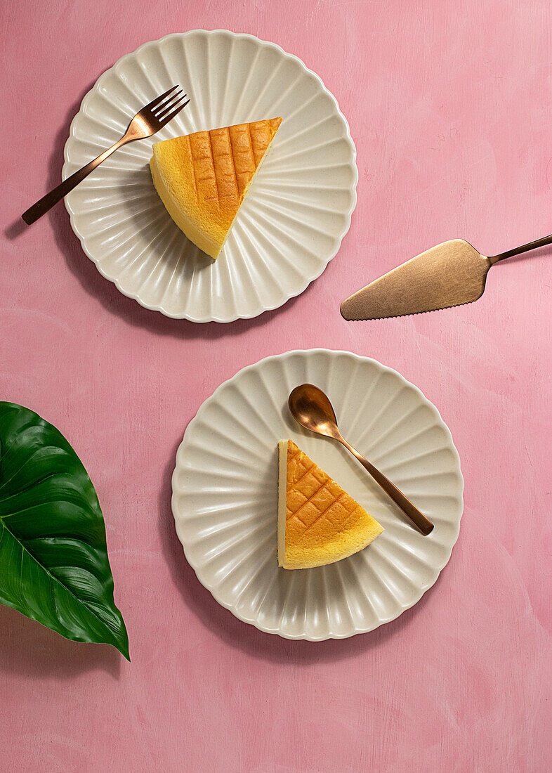 Draufsicht auf Teller mit Scheiben von leckerem japanischem Käsekuchen und Besteck neben Spatel und grünem Pflanzenblatt auf rosa Hintergrund