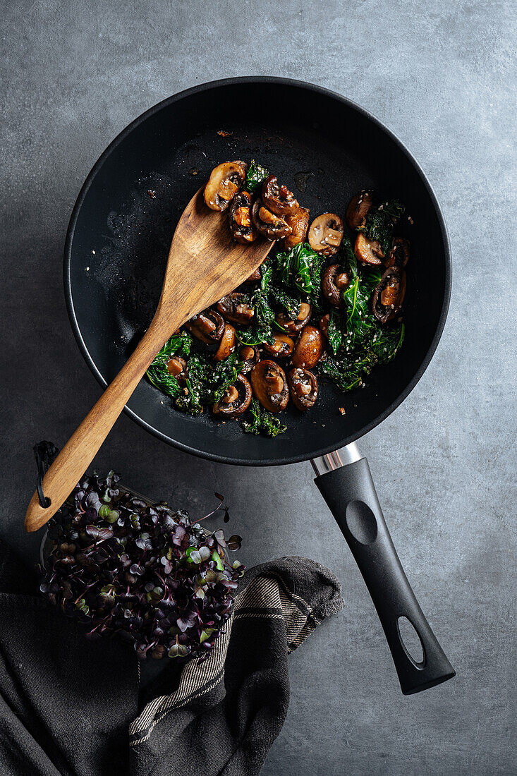 From above wooden spatula putting tasty sauteed mushrooms with greens from frying pan on plate