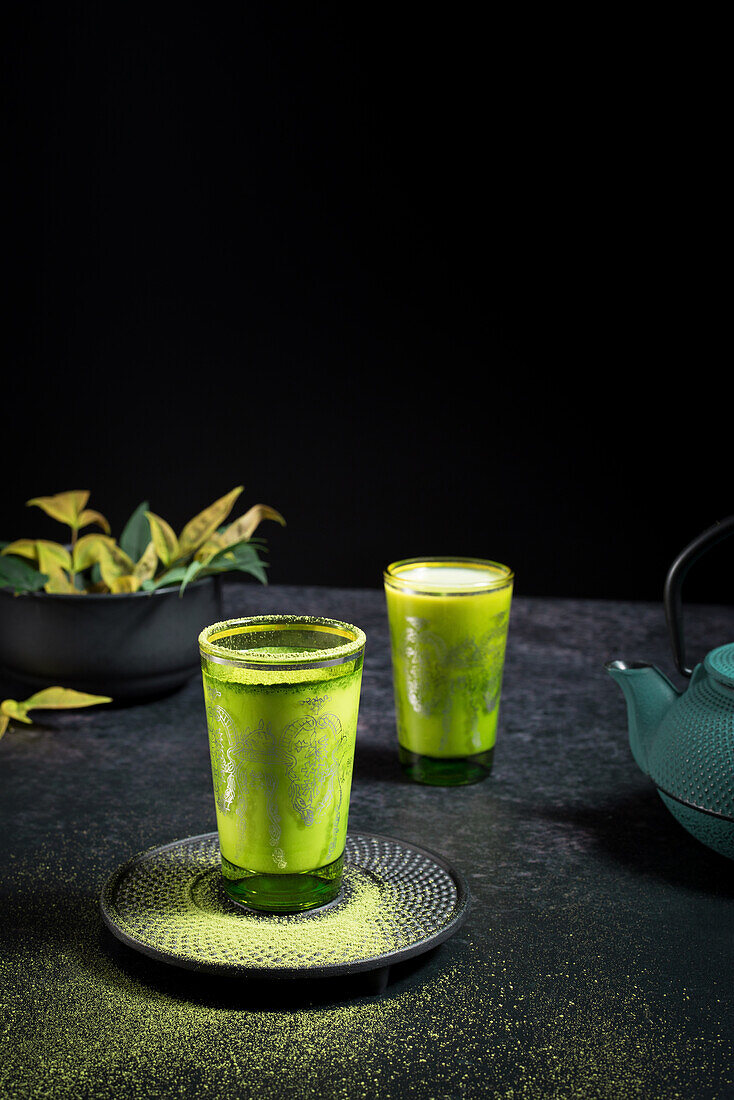 Still life composition with traditional oriental matcha tea served in glasses cups with metal ornamental decor on table with ceramic bowls and fresh green leaves against black background