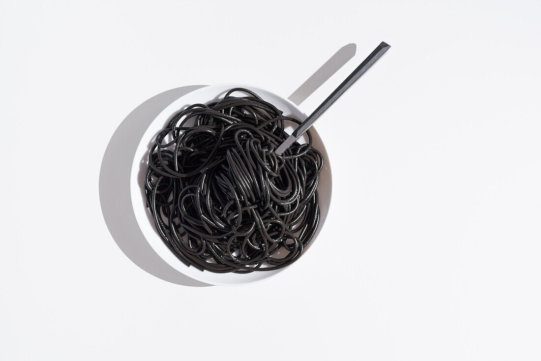 Blick von oben auf eine rostfreie Gabel in einer vollen Schüssel mit schwarzen Spaghetti mit Tintenfischtinte auf einem weißen Tisch im Studio