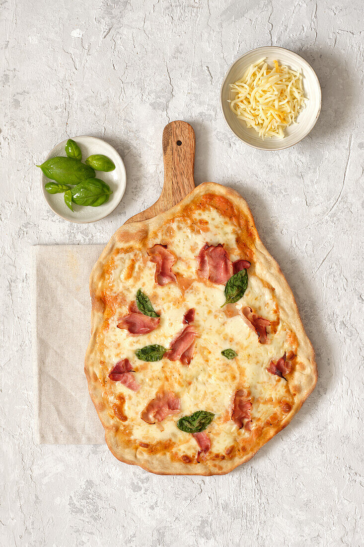 Blick von oben auf eine leckere hausgemachte Pizza mit Basilikum und Schinken, serviert auf einem hölzernen Schneidebrett auf dem Tisch