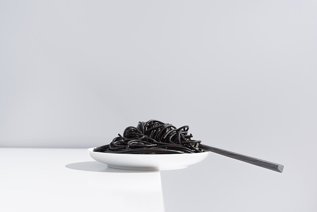 Stainless fork in full bowl of black spaghetti with cuttlefish ink on white table in studio on gray background