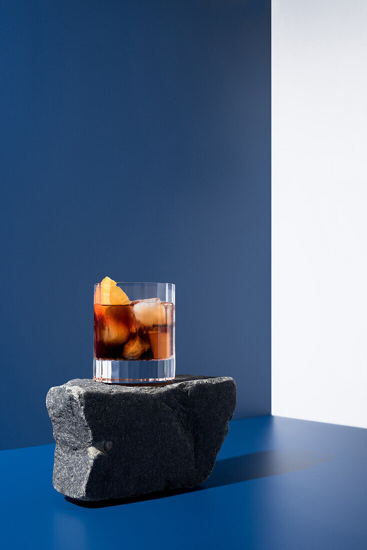 Glass of alcoholic cocktail made with red vermouth served with orange slices on stone in light studio with blue and white wall