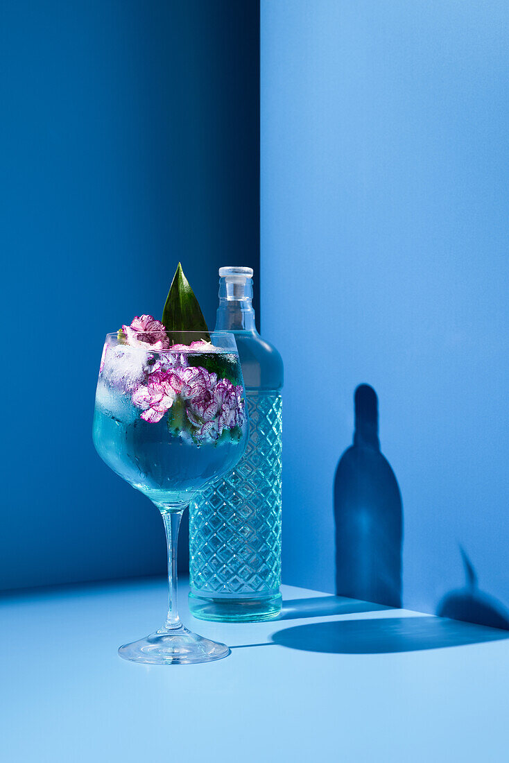Transparent glass bottle of alcohol drink placed on table near fresh cocktail with ice and flowers against blue background