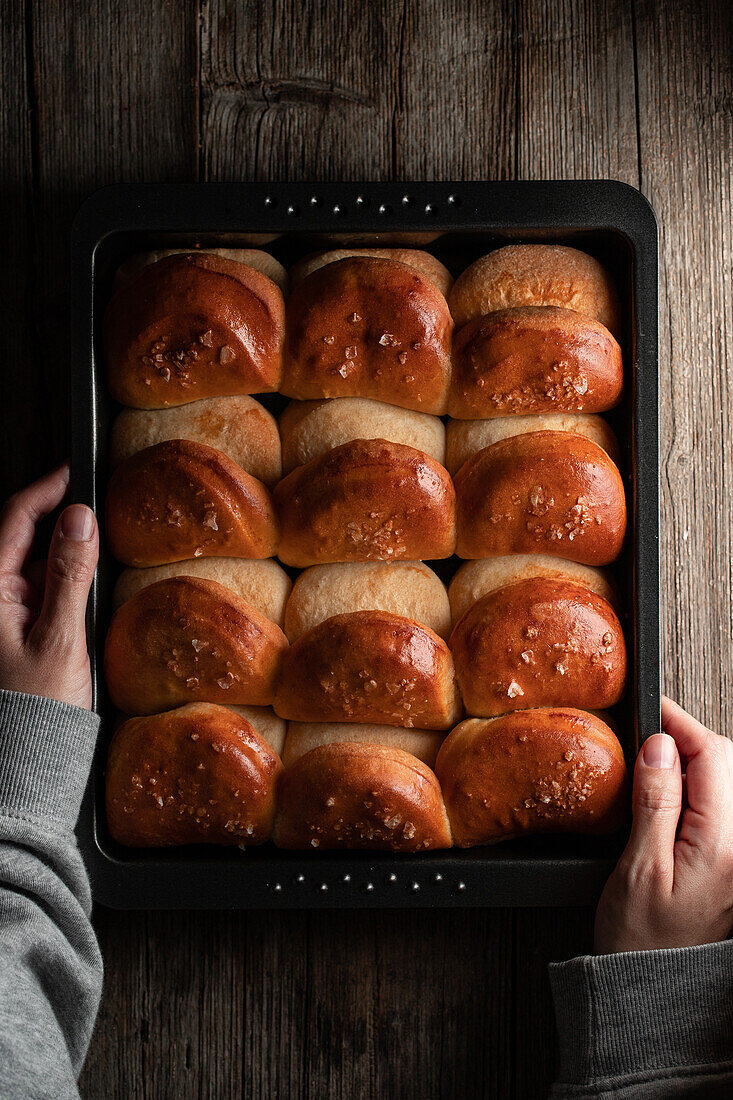 Blick von oben auf einen gesichtslosen Erntekoch, der eine Backform mit appetitlichen weichen Brötchen auf einem Holztisch hält