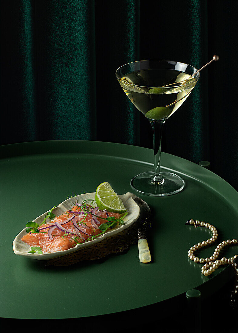 From above of appetizing smoked salmon slices served on plate with lime and onion and placed on round table with glass of vermouth with olive near elegant pearl necklace