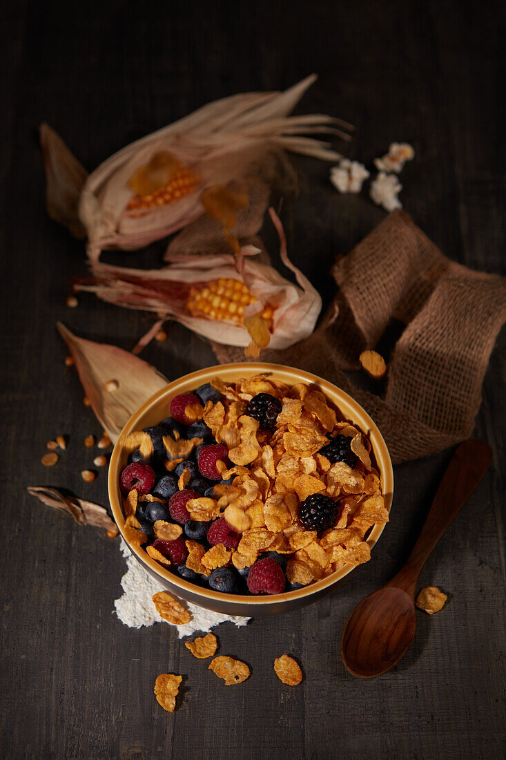 From above tasty cornflakes with fresh forest berries served in bowl near burlap fabric during breakfast in dark room