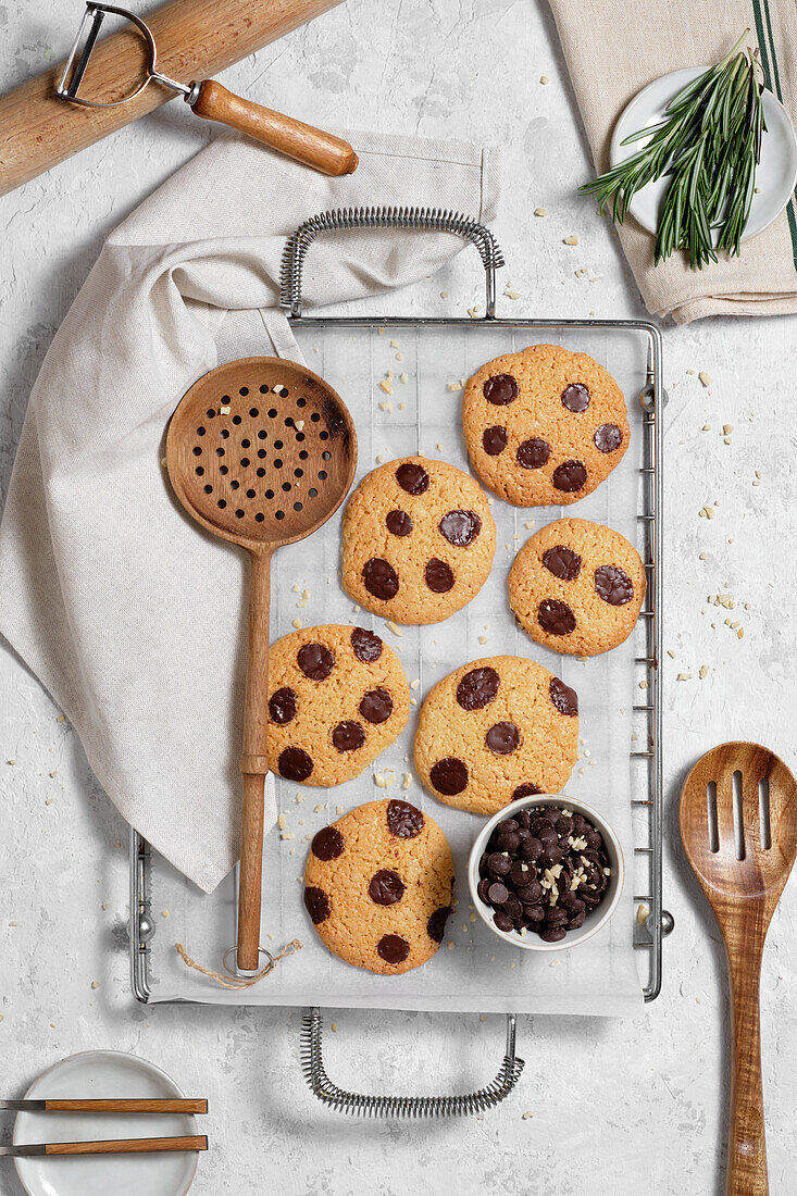 Top view of freshly baked sweet cookies with chocolate chips on metal grid placed on table with various kitchen tools and green rosemary branches