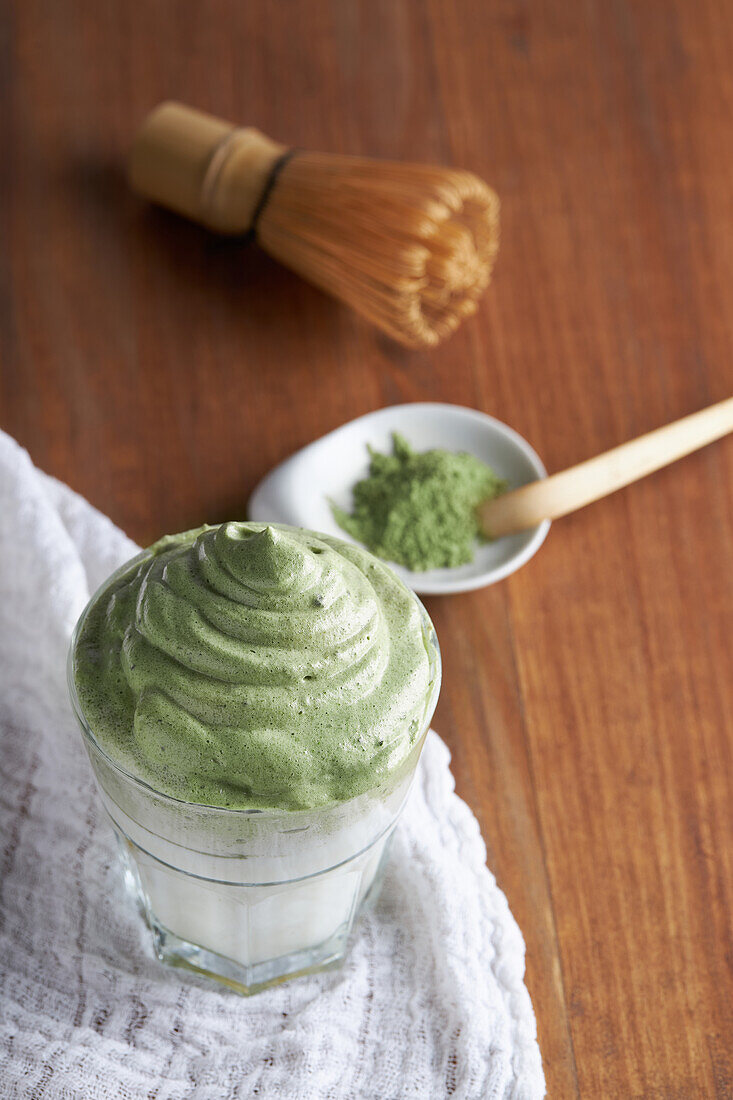 From above glass of fresh dalgona coffee with matcha and Japanese tea ceremony accessories consisting of bamboo whisk called chasen and traditional scoop named chashaku placed on wooden table