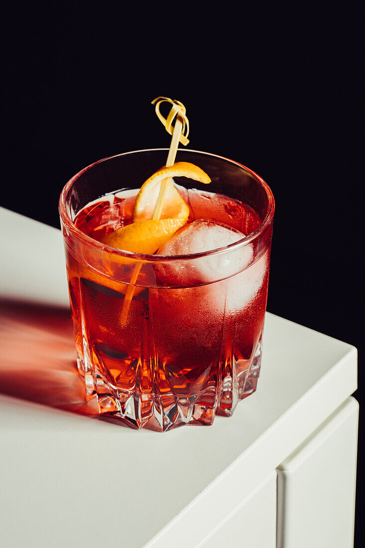 Glass of bitter alcoholic Negroni cocktail served with ice and orange peel on white surface