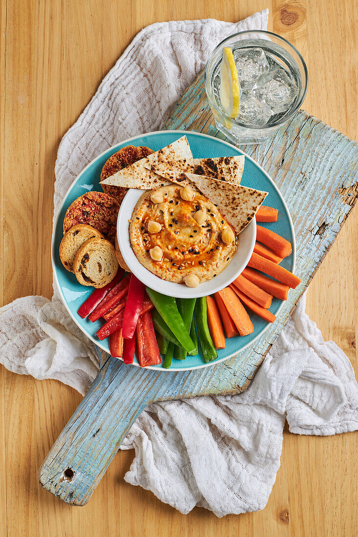 Draufsicht auf leckeren Hummus mit Tortilla-Chips, serviert auf einem Teller mit Croutons und verschiedenen Gemüsesorten auf einem rustikalen Schneidebrett neben Gläsern mit kalter Limonade auf einem Holztisch mit Serviette