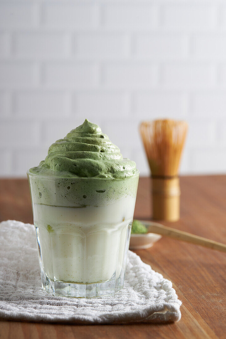 Glass of fresh dalgona coffee with matcha and Japanese tea ceremony accessories consisting of bamboo whisk called chasen and traditional scoop named chashaku placed on wooden table