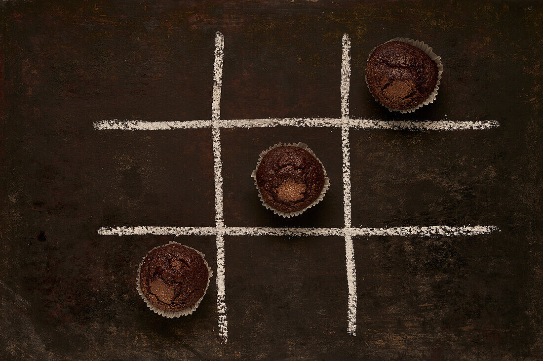 Draufsicht auf ein essbares Tic-Tac-Toe-Spiel mit gebackenen Schokoladenmuffins, die Noughts darstellen, auf schwarzem Hintergrund