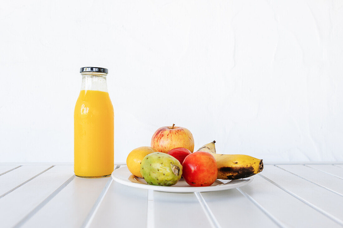 Flasche mit frischem Orangensaft auf weißem Tisch neben Teller mit verschiedenen bunten reifen Früchten im Tageslicht