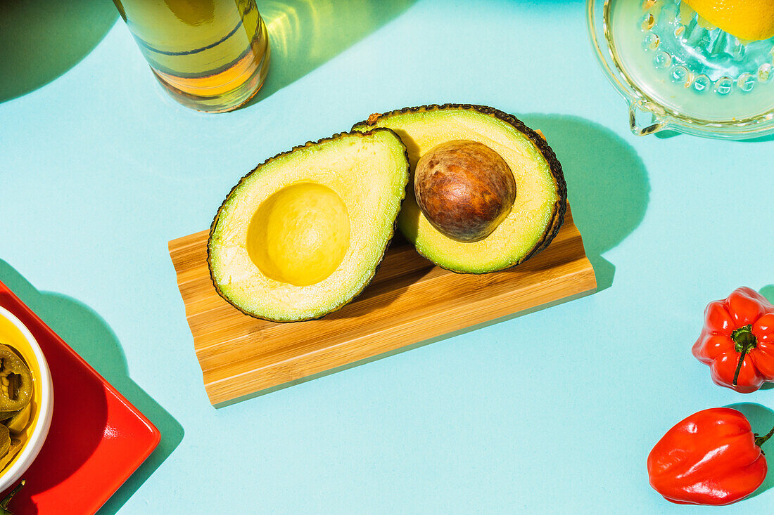 Draufsicht auf eine halbierte reife Avocado, die auf einem Holzbrett in der Nähe von Habanero-Pfeffer auf blauem Hintergrund liegt