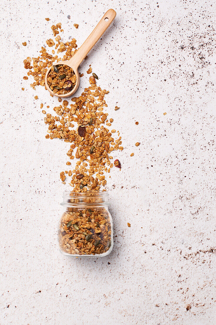 From above crispy granola spilled from glass jar near wooden spoon on light table