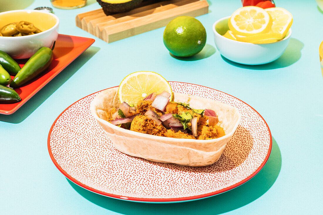 Portion leckeres mexikanisches Brathähnchen mit Zwiebeln und Kräutern, serviert mit Zitrone in Tortillaschale auf Teller auf blauem Tisch