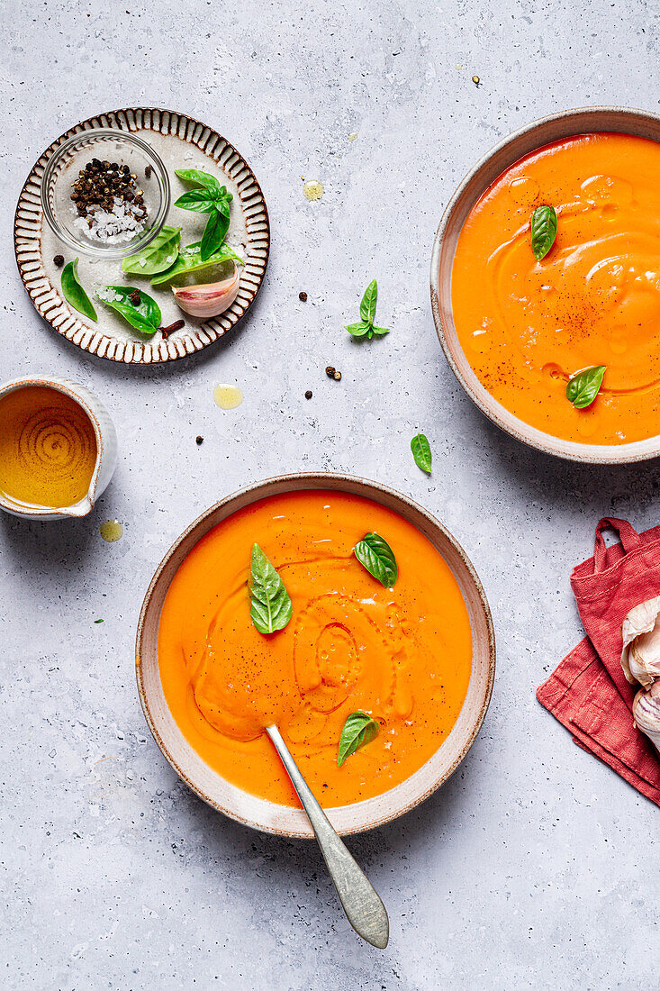 Top view of bowls with tomato soup decorated with basil and placed on table near sauce and salt with garlic