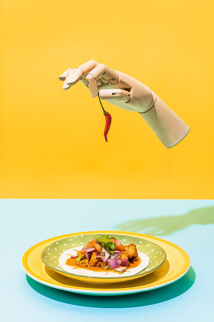 Artificial hand with red hot chili pepper levitating over plate with Mexican chicken salad on blue and yellow background