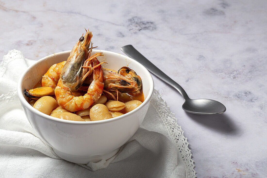 From above bowl with typical stew of beans with prawns, shrimps and mussels on a lace tablecloth close to a spoon and on a stone table