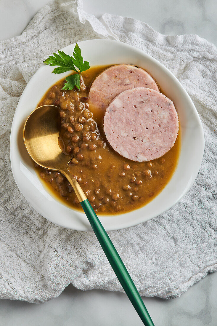 Top view of spoon placed in bowl of yummy lentil soup served with slices of sausage and leaf of parsley on marble table and gray napkin