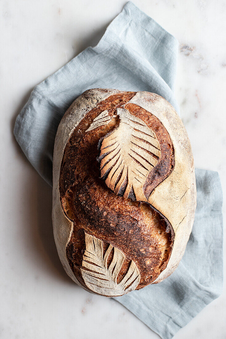 Draufsicht auf einen frisch gebackenen Laib Brot mit Blattornament auf einer Serviette auf weißem Hintergrund in der Küche
