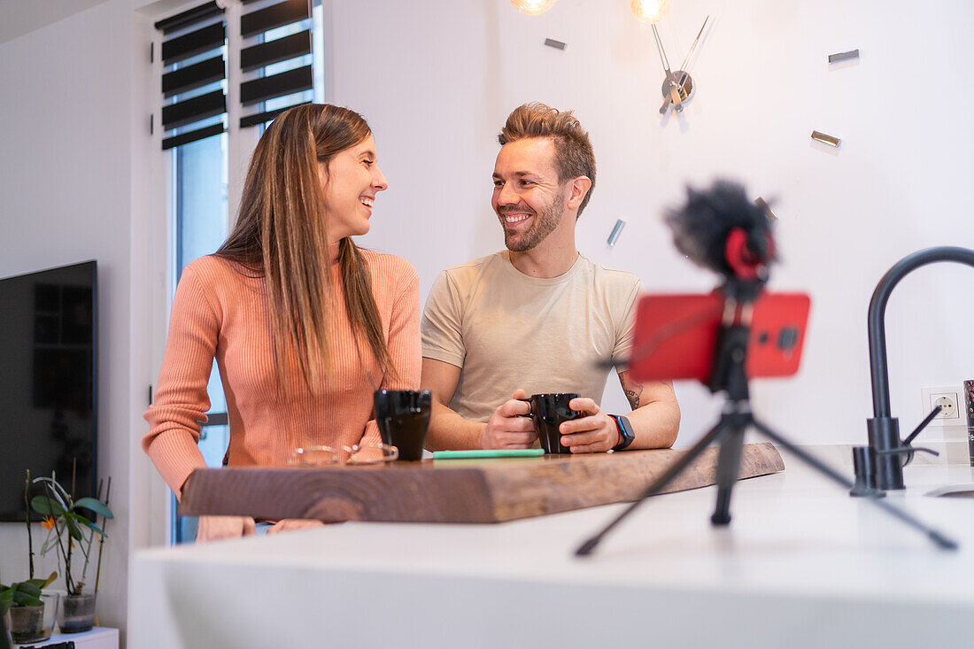 Positives Bloggerpaar, das einen Vlog mit Smartphone und Mikrofon aufnimmt, während es zu Hause am Küchentisch sitzt und eine Tasse Kaffee trinkt