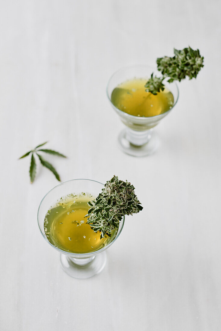 Von oben kleine Gläser mit medizinischem Marihuana-Tee und grünen Kräutern auf weißer Fläche neben Glasgefäß platziert