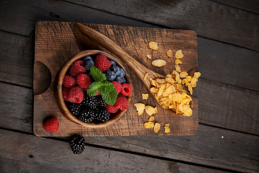 Top view of spoon with cornflakes near bowl with heap of various ripe berries on wooden chopping board in kitchen