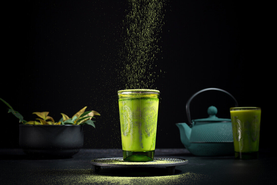 Still life composition with traditional oriental matcha tea served in glass cup with metal ornamental decor on table with ceramic bowls and fresh green leaves against black background