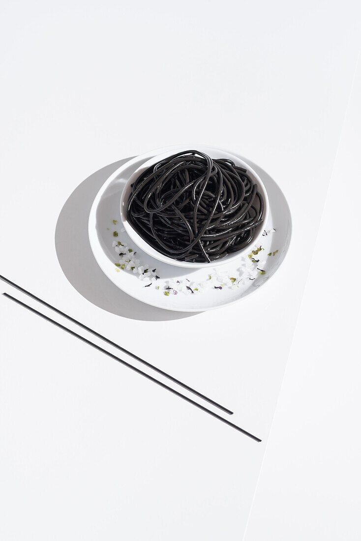 Blick von oben auf eine Keramikschale mit leckeren Spaghetti mit schwarzer Tintenfischtinte und Stäbchen auf weißem Hintergrund