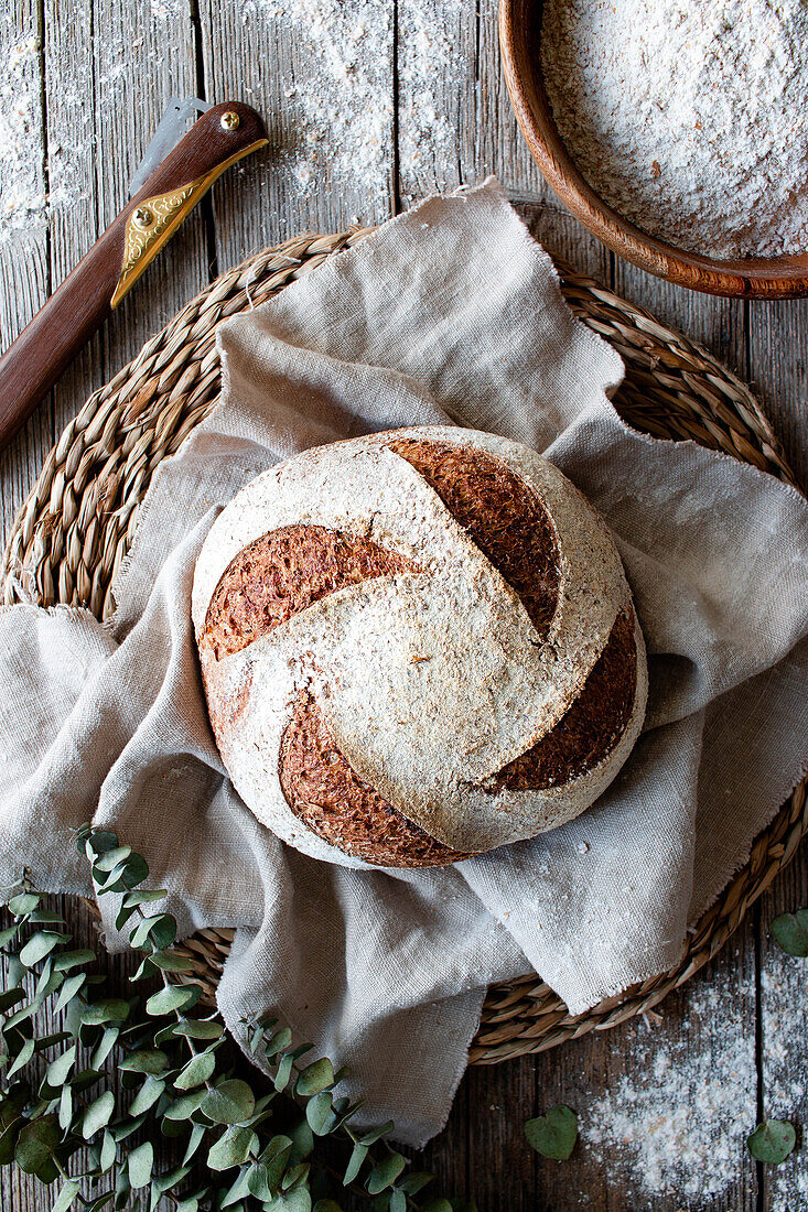 Von oben hausgemachtes frisches Dinkelsauerteigbrot auf Weidenständer mit Tuch auf Holztisch mit Mehl bestreut