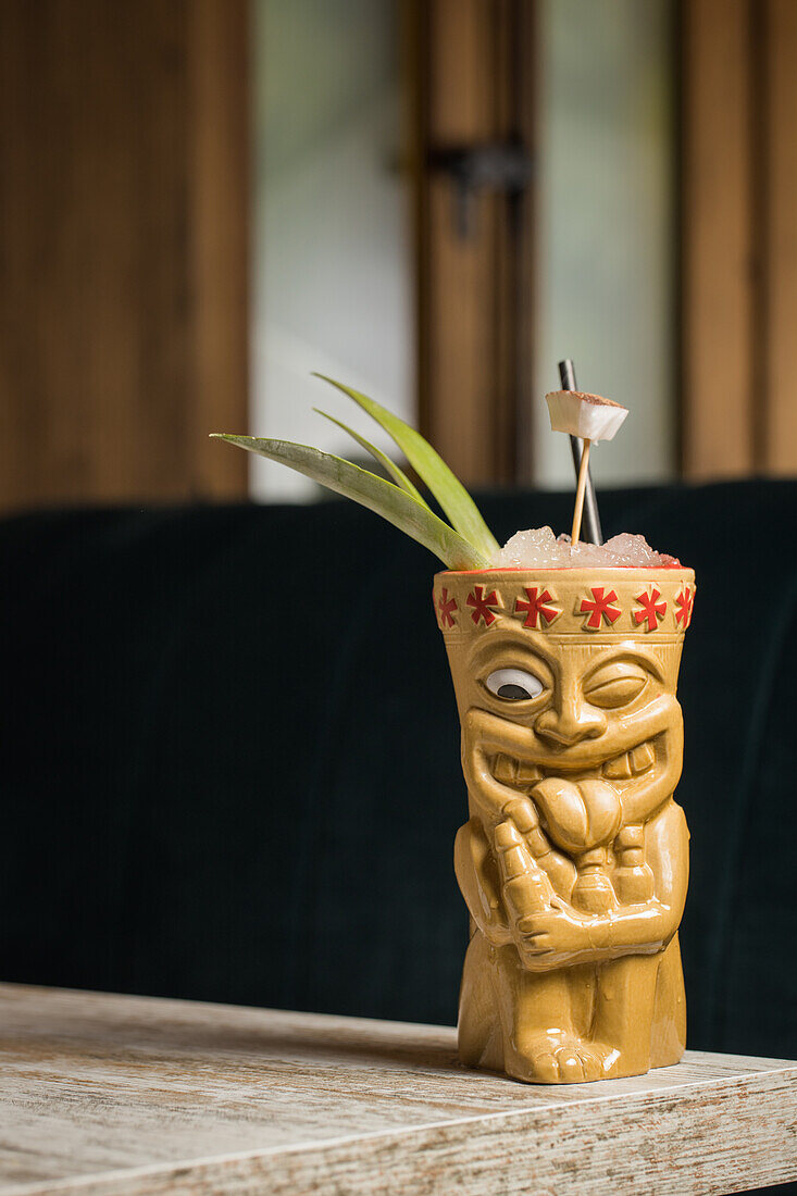 Polynesian tiki cup of cold alcohol beverage decorated with straw and green pineapple leaves placed against on wooden table
