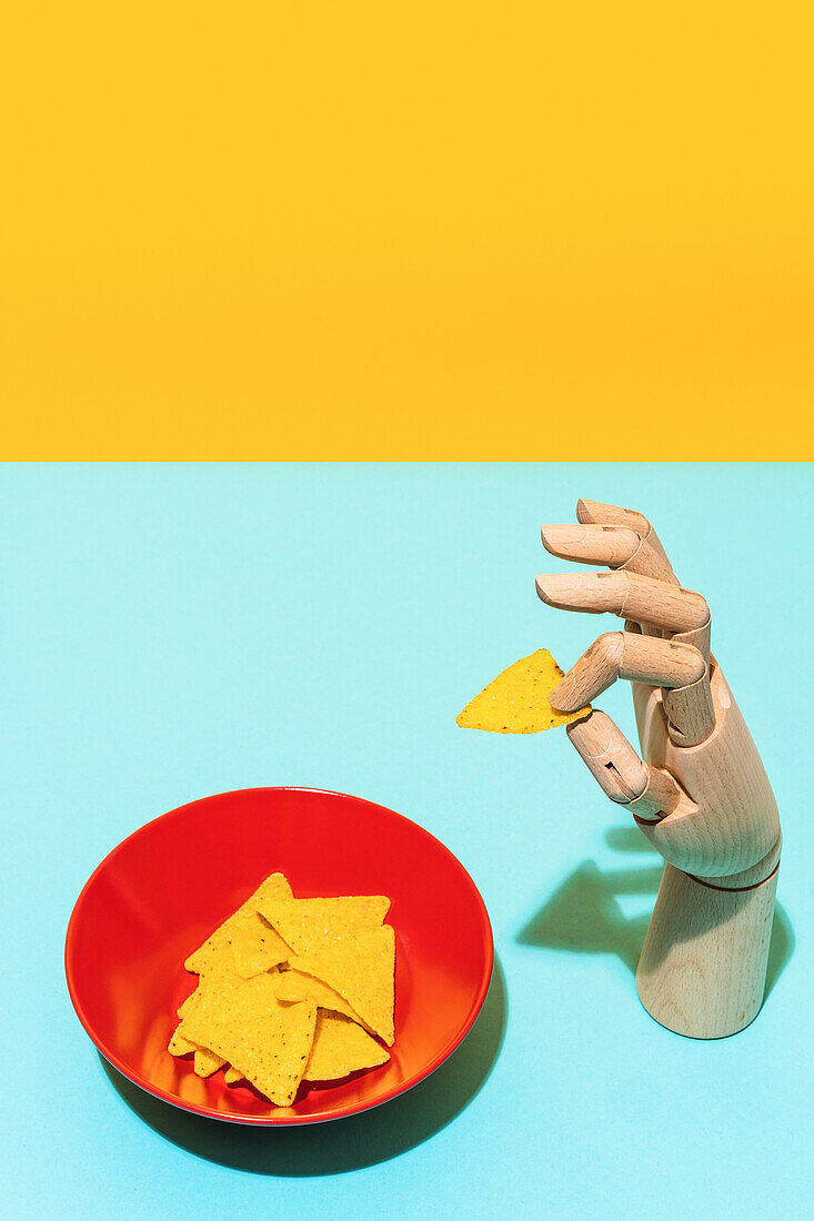 From above artificial hand holding tortilla chip placed near bowl with tortilla chips for Mexican cuisine dish on yellow and blue background