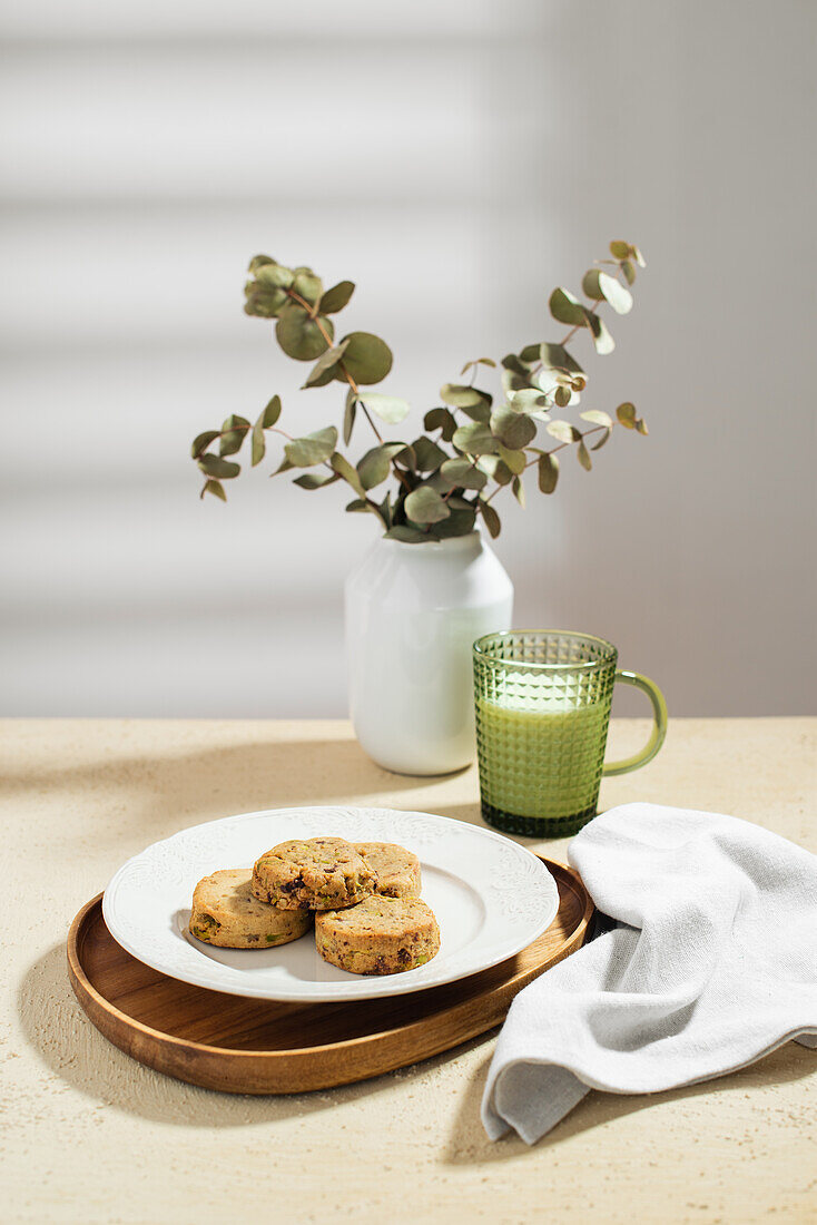 Plate with stack of tasty biscuits and mug of fresh milk placed on table near napkin and vase with twigs in morning