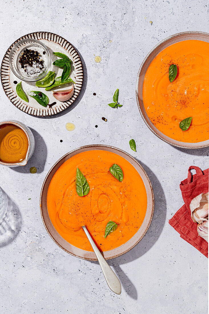 Top view of bowls with tomato soup decorated with basil and placed on table near sauce and salt with garlic