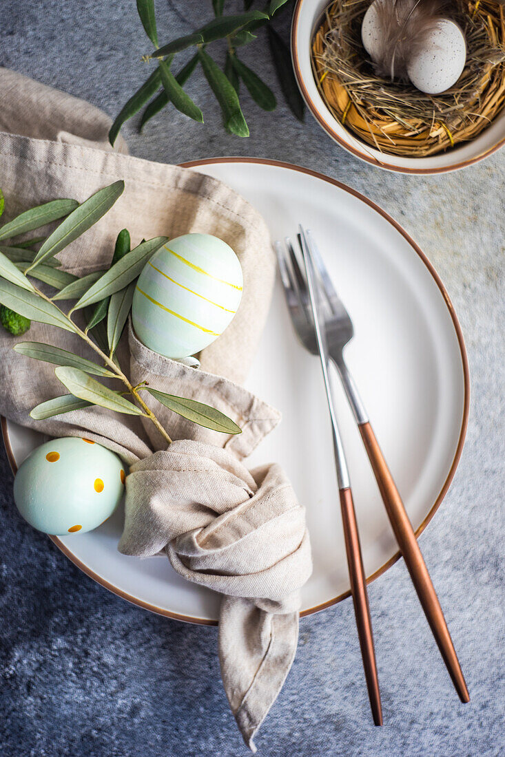 From above cutlery set for Easter dinner with olive tree branches and eggs on a bird nest on a concrete background