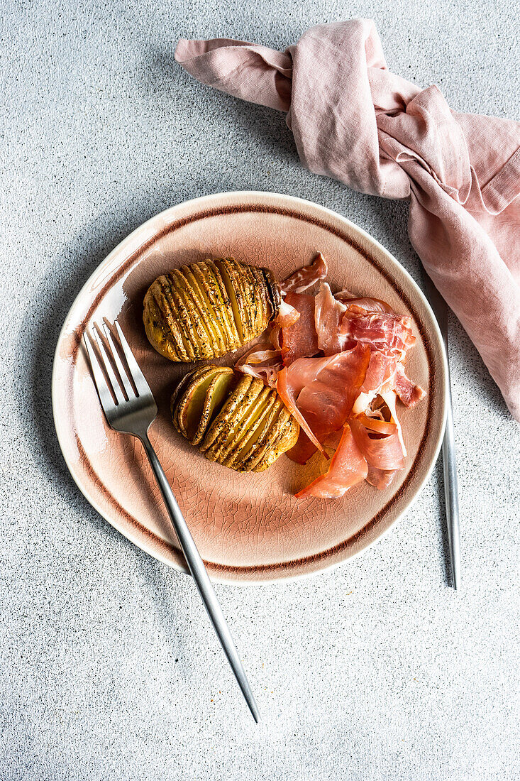 Von oben gebackene gewürzte Hasselback-Kartoffeln mit Speckscheiben, serviert in einer rosa Keramikschüssel auf Betonhintergrund