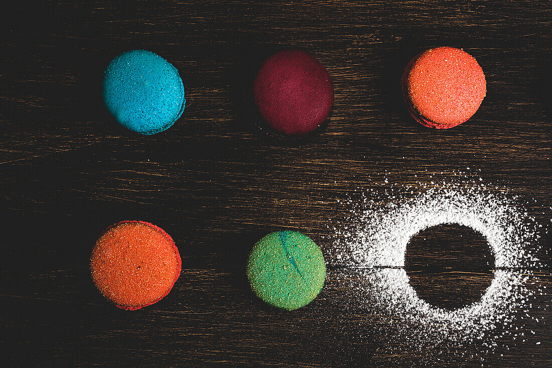 From above colorful French macaroons with various fillings placed on wooden table near powder sugar in dark background