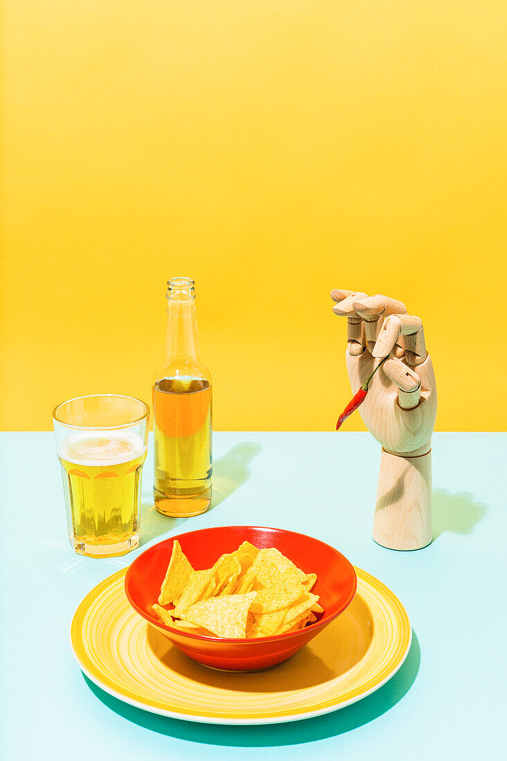 Von oben Vergrößerungsglas und künstliche Hand hält scharfe Paprika und Schüssel mit Tortilla-Chips für mexikanische Küche Gericht auf gelben und blauen Hintergrund